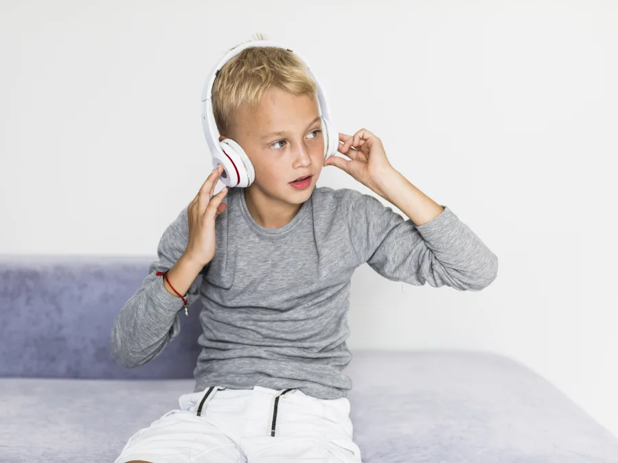 Young child wearing white over-ear headphones and touching the earcups while sitting indoors and looking to the side.